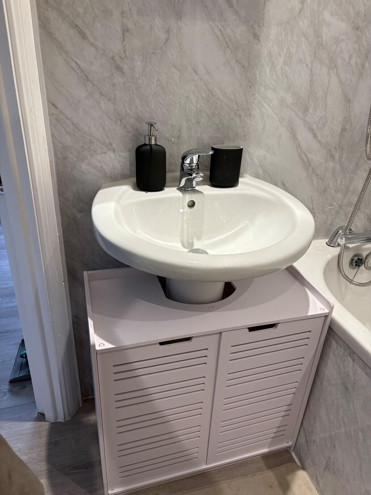 A bathroom with a white sink, a mirror, a handheld soap dispenser, and a shower.