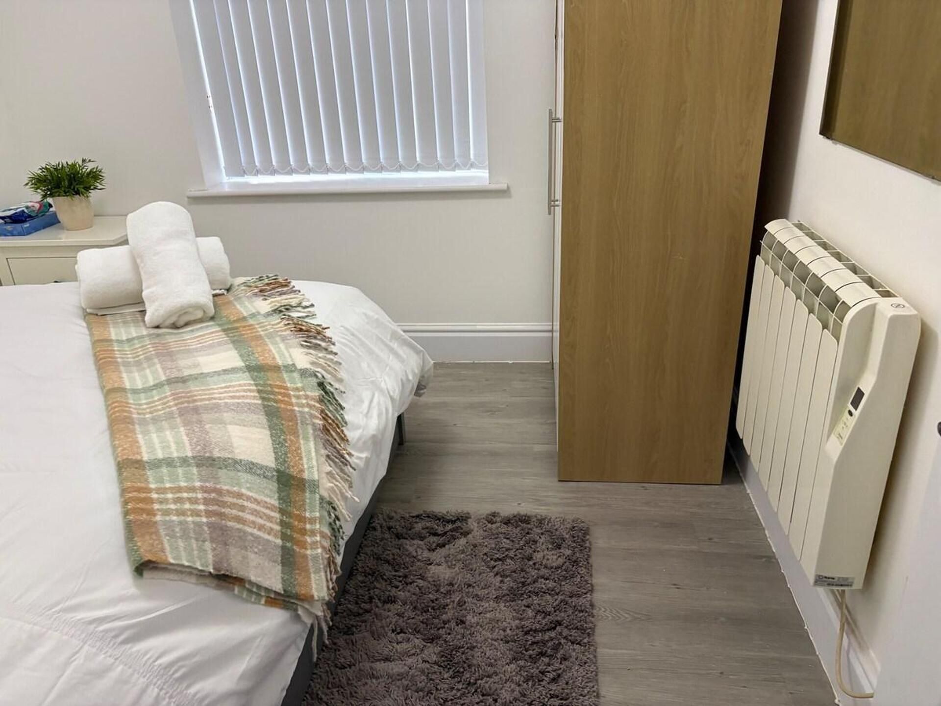 Ein Schlafzimmer mit Bett, Heizkörper, Fenster mit Jalousien, Tür und einer kleinen Pflanze auf einem Nachttisch.