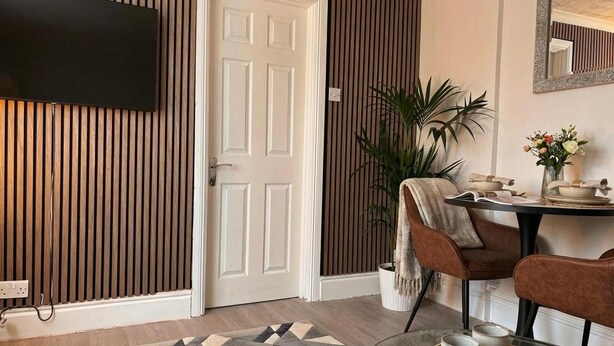A room with a wooden slat wall, a white door, a television mounted above it, a potted plant, a table with chairs, and a patterned rug. - Near Etihad/coop Live - Parking - Wifi - Sleeps 4 (Manchester)