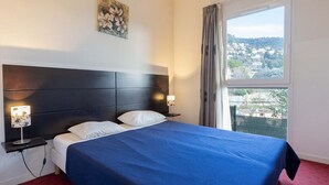 A hotel room with a bed, two bedside tables, a nightstand, a window with a view, and a painting on the wall. - Air-conditioned flat for 4 people, with a swimming pool and balcony (Grasse)