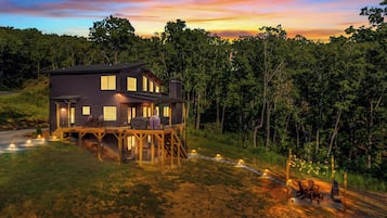A two-story house with a deck, surrounded by trees and a sunset sky.