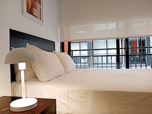 A hotel room with a bed, a nightstand, a lamp, a window with a view of the outside, and a painting on the wall. - Travelers Smart Suites - TAJ 97 (Bogotá)