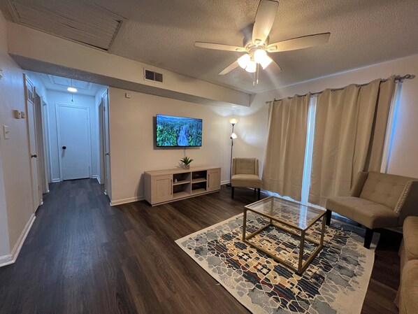 A living room with a ceiling fan, a flat-screen TV, a beige sofa, a coffee table, and a patterned rug.