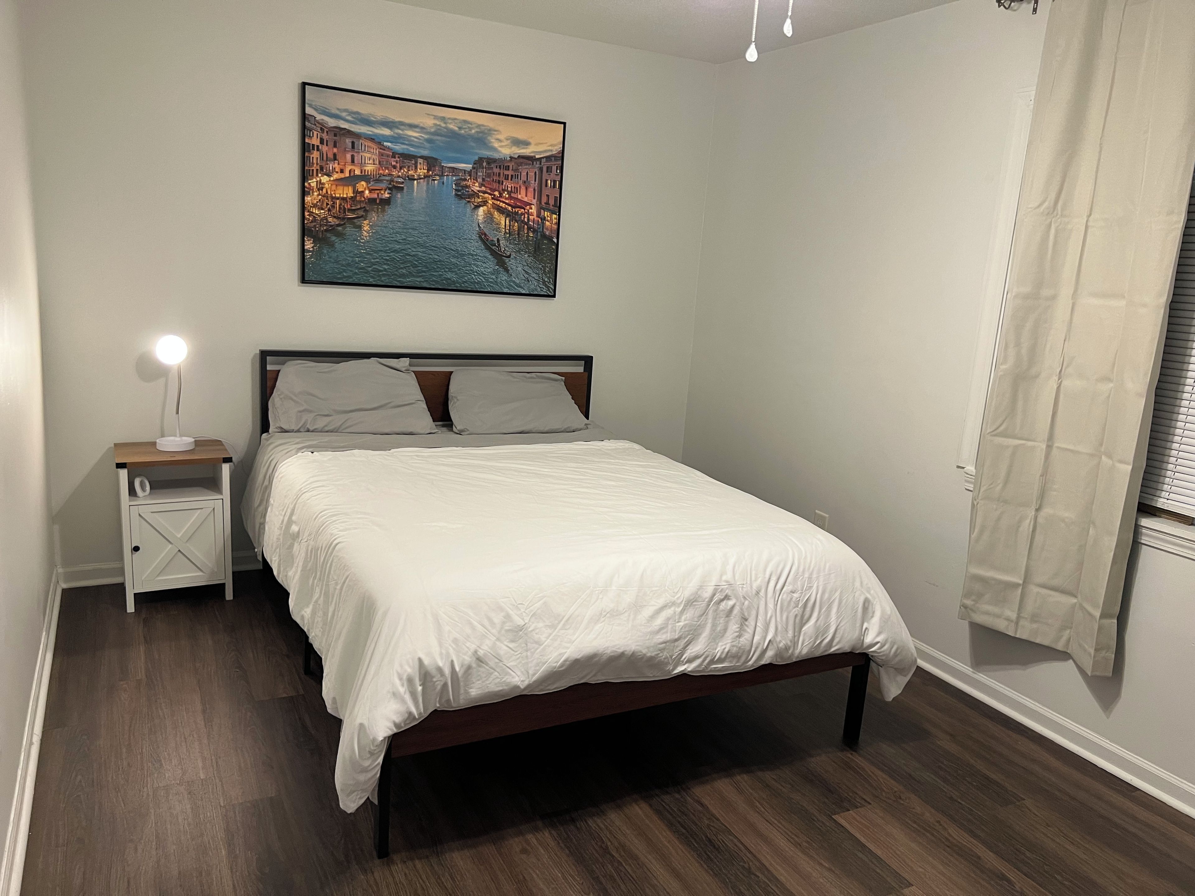 Un dormitorio con cama, mesita de noche con lámpara, un cuadro de un canal y una ventana con cortinas.