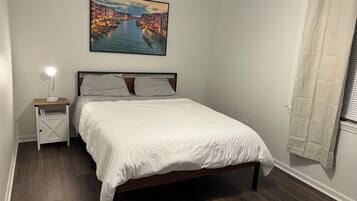 Un dormitorio con cama, mesita de noche con lámpara, un cuadro de un canal y una ventana con cortinas.