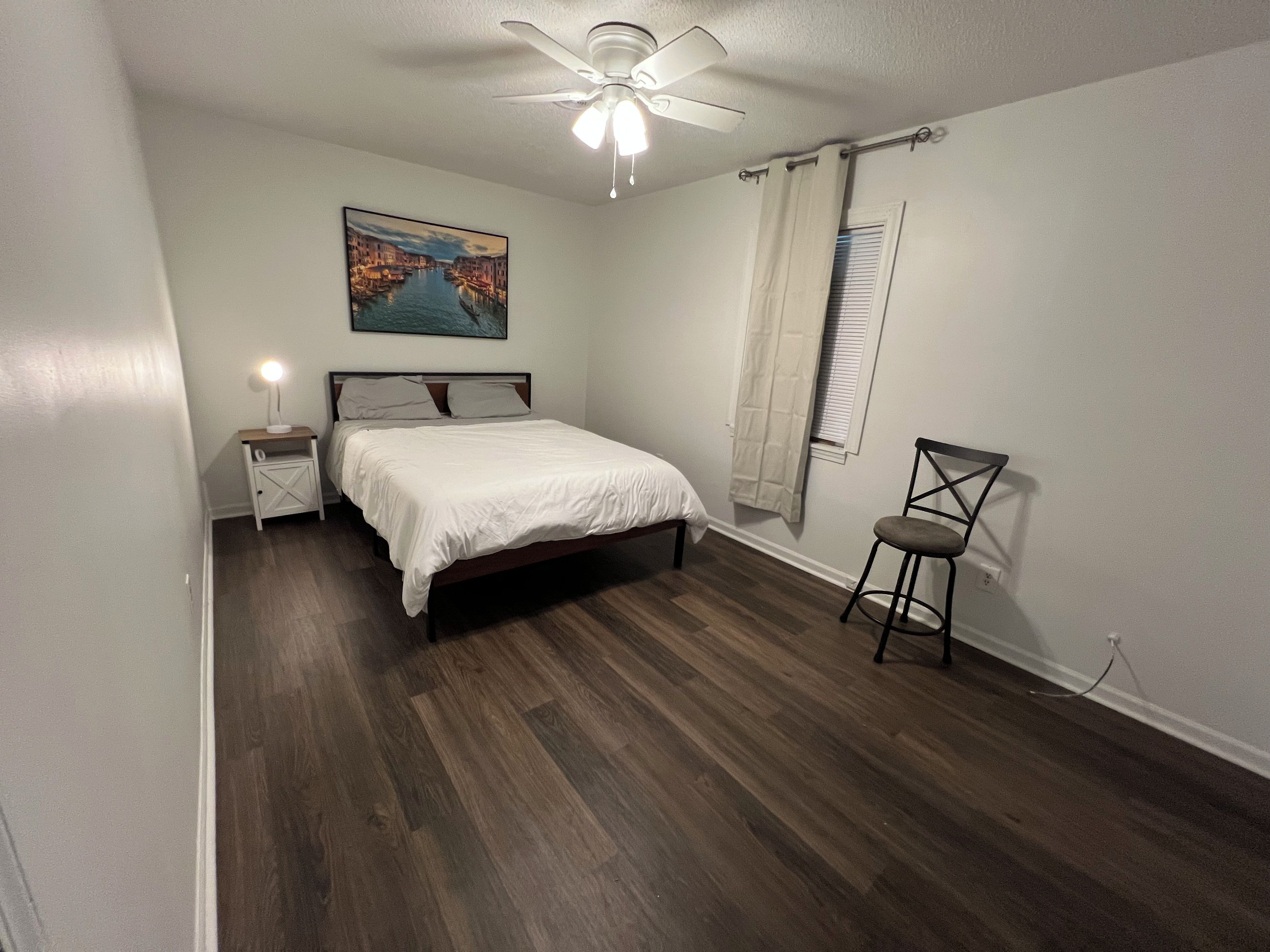 Una camera da letto con un letto, un comodino, una sedia, una ventola a soffitto, una finestra con tende e un quadro appeso al muro.