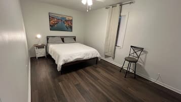 Una camera da letto con un letto, un comodino, una sedia, una ventola a soffitto, una finestra con tende e un quadro appeso al muro.