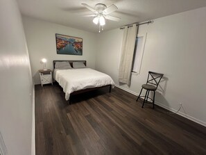 A bedroom with a bed, a nightstand, a chair, a ceiling fan, a window with curtains, and a painting on the wall.