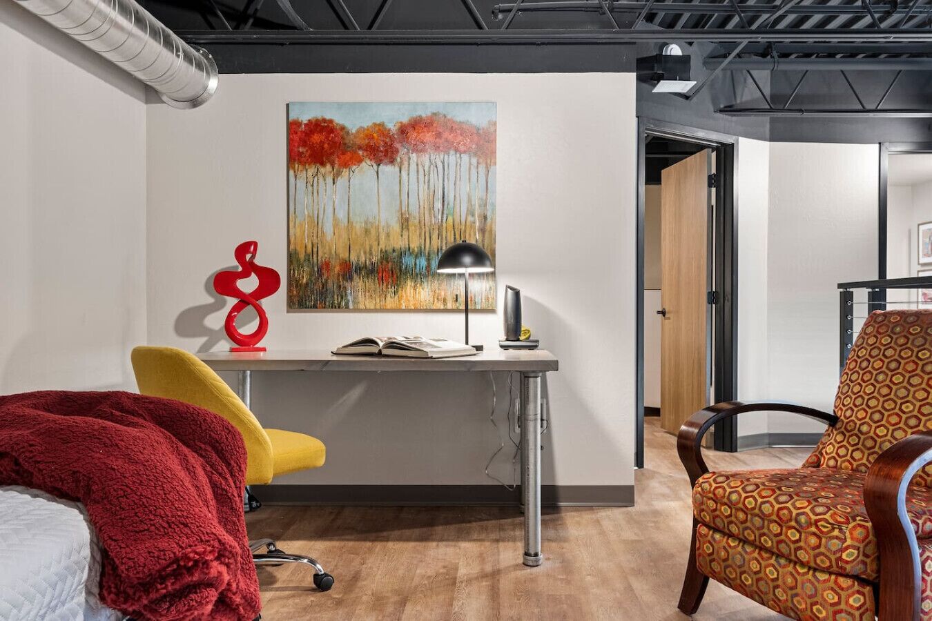 A modern office space with a desk, a yellow chair, a red abstract sculpture, and a patterned armchair.