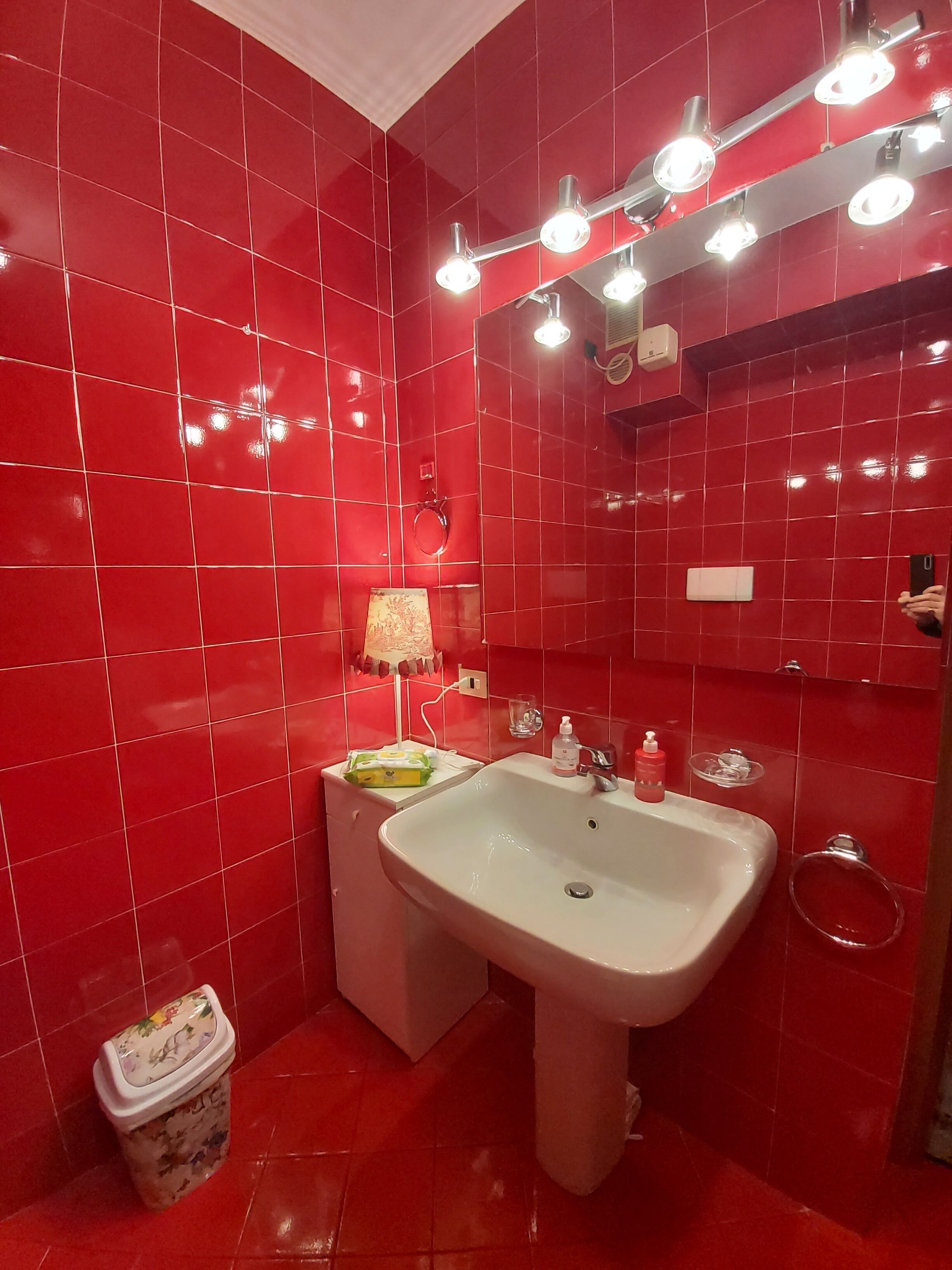 A bathroom with red tiles, a sink, a mirror, and a trash bin.