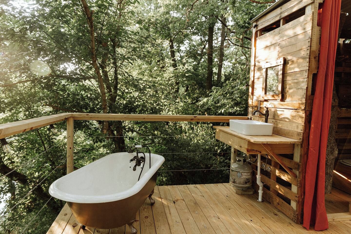 A wooden deck with a freestanding bathtub and a sink, surrounded by trees.