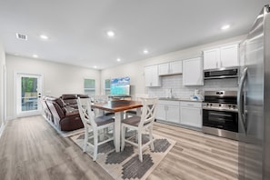 A modern kitchen with white cabinets, stainless steel appliances, and a dining area with a leather sofa and white chairs. - Marigold House (Foley)