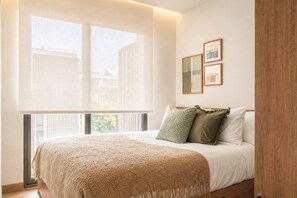 A bedroom with a large window, a bed with a beige blanket, and two framed pictures on the wall. - Luxury 2br in Laguna - Golf view - skypark pool (Choeng Thale)
