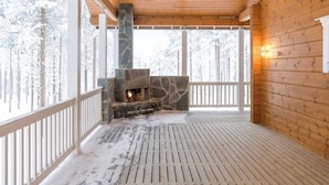 A wooden cabin with a stone fireplace, a wooden deck, and a view of snow-covered trees. - Charming Lapland Villa With Mountain Views, Private Sauna and Outdoor Hottub (Pelkosenniemi)