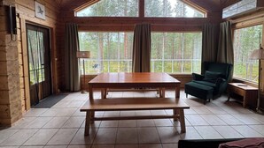 A wooden cabin interior with a large table, two windows with curtains, a chair, and a lamp. - Charming Lapland Villa With Mountain Views, Private Sauna and Outdoor Hottub (Pelkosenniemi)