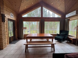 A wooden cabin interior with a large table, two windows with curtains, a chair, and a lamp. - Charming Lapland Villa With Mountain Views and Private Sauna (Pelkosenniemi)