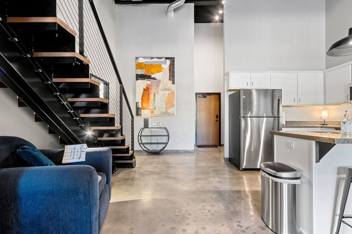 A modern kitchen with stainless steel appliances, a dining area with a blue sofa, and a staircase with black railings.
