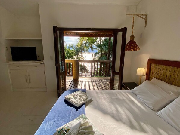 Un dormitorio con cama, televisión en un mueble, balcón con vista a palmeras y una lámpara colgante.