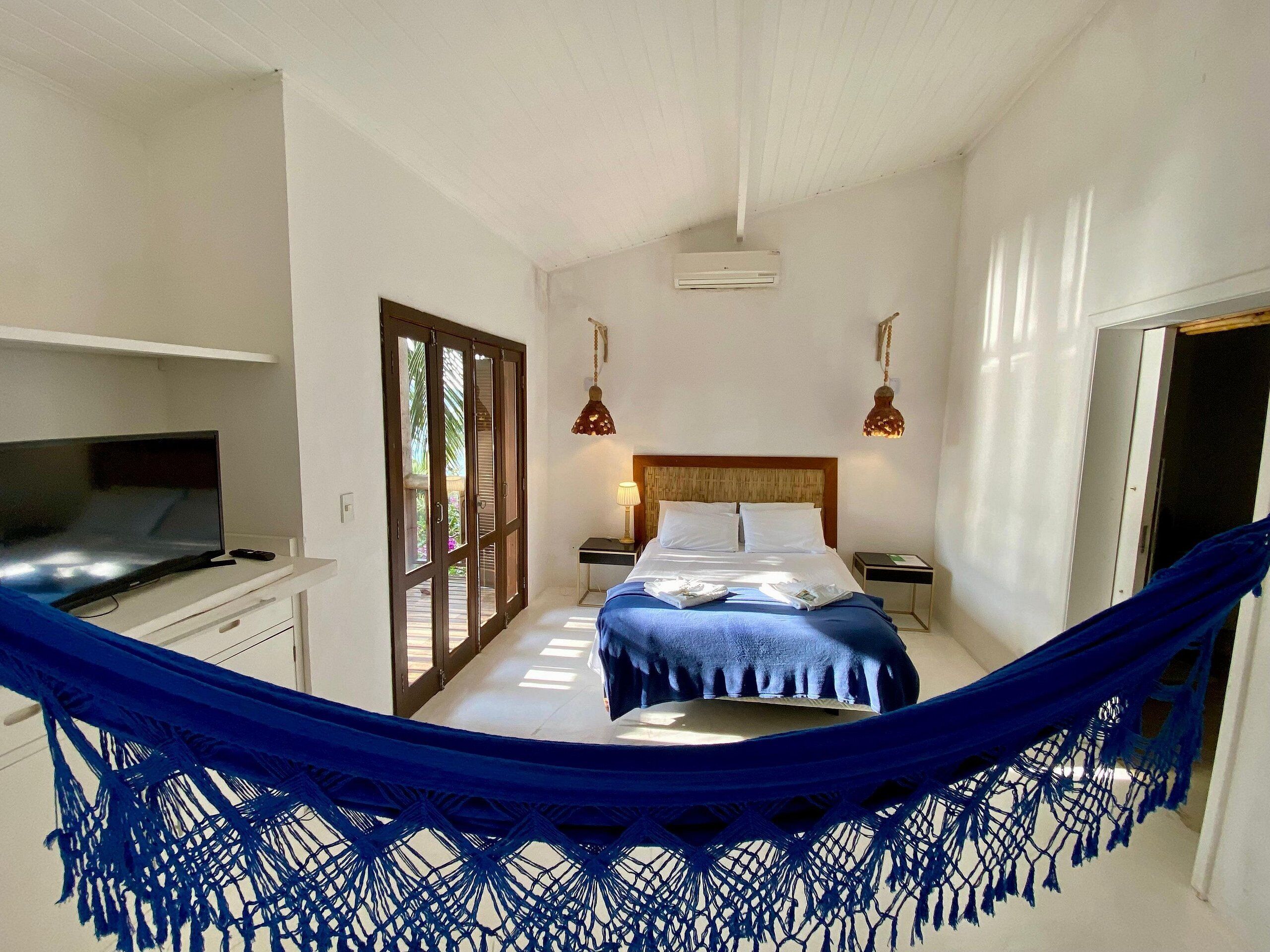 A bedroom with a bed, a desk with a TV, a hammock, and a door leading to another room.