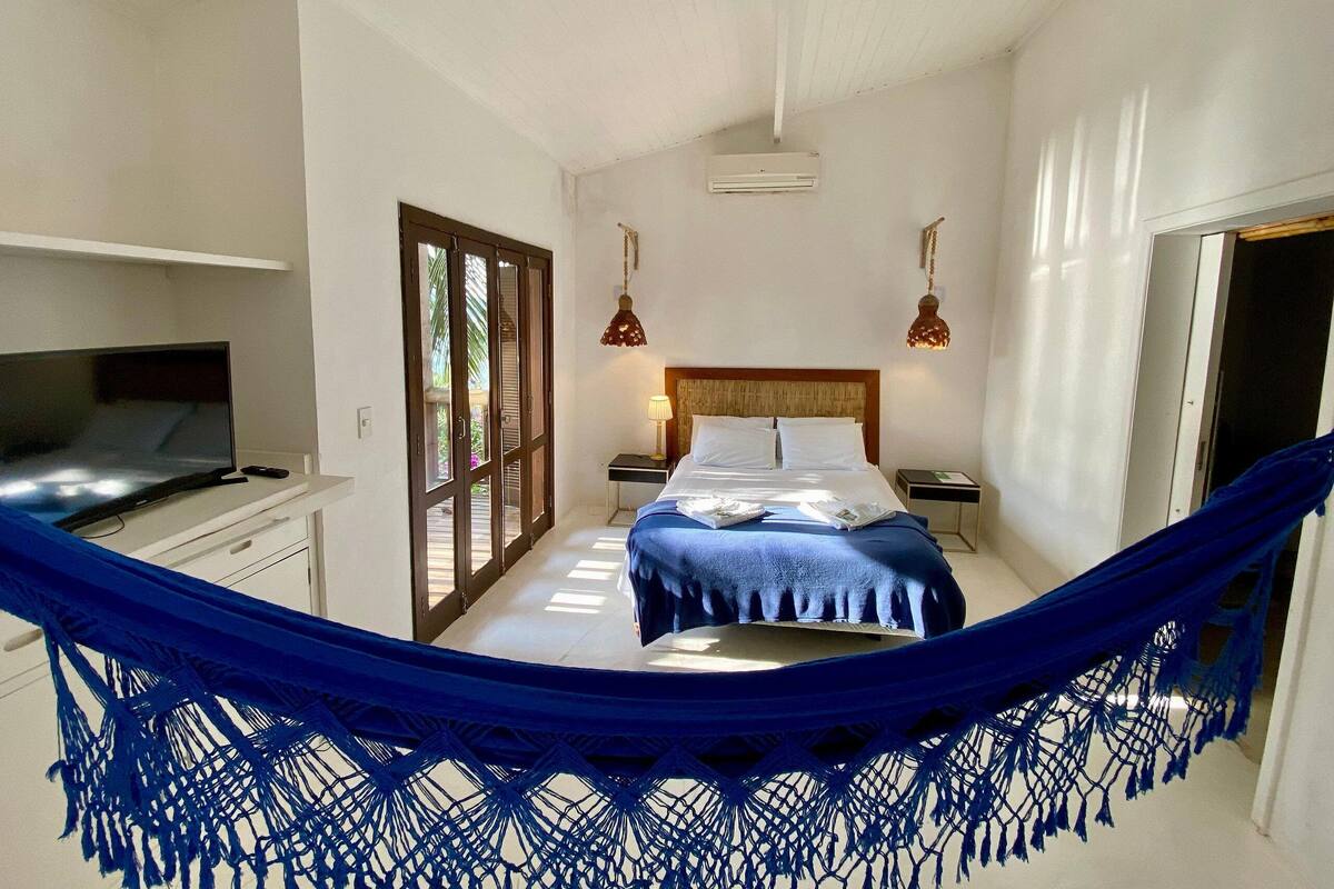 A bedroom with a bed, a desk with a TV, a hammock, and a door leading to another room.