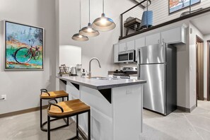 Una cocina moderna con una isla central, electrodomésticos de acero inoxidable, iluminación colgante y un cuadro en la pared.