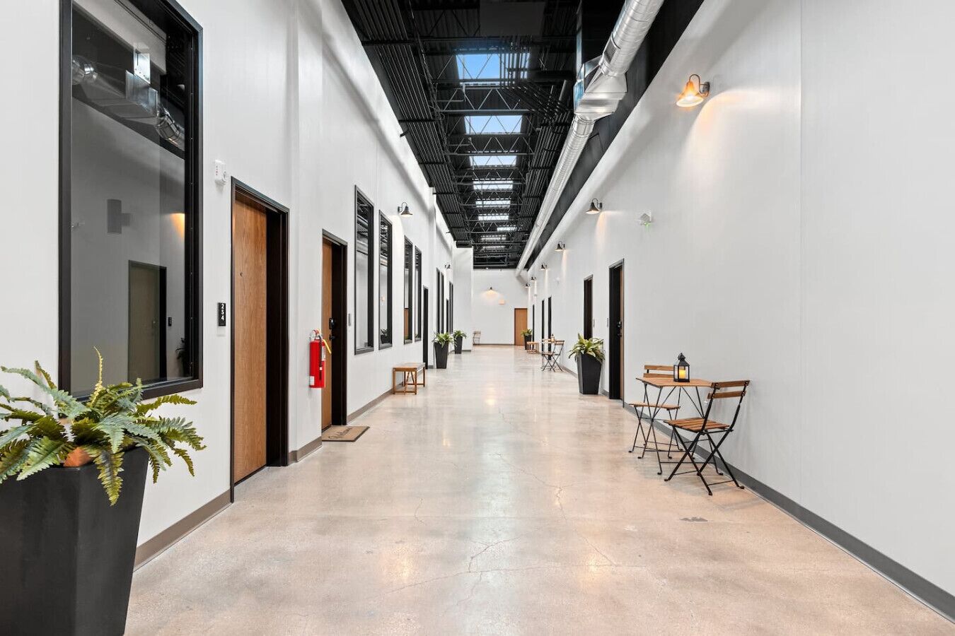 Un long couloir étroit aux murs blancs, aux portes à encadrement noir et au sol en béton. Des plantes en pot et une petite table avec des chaises sont disposées le long du côté droit.