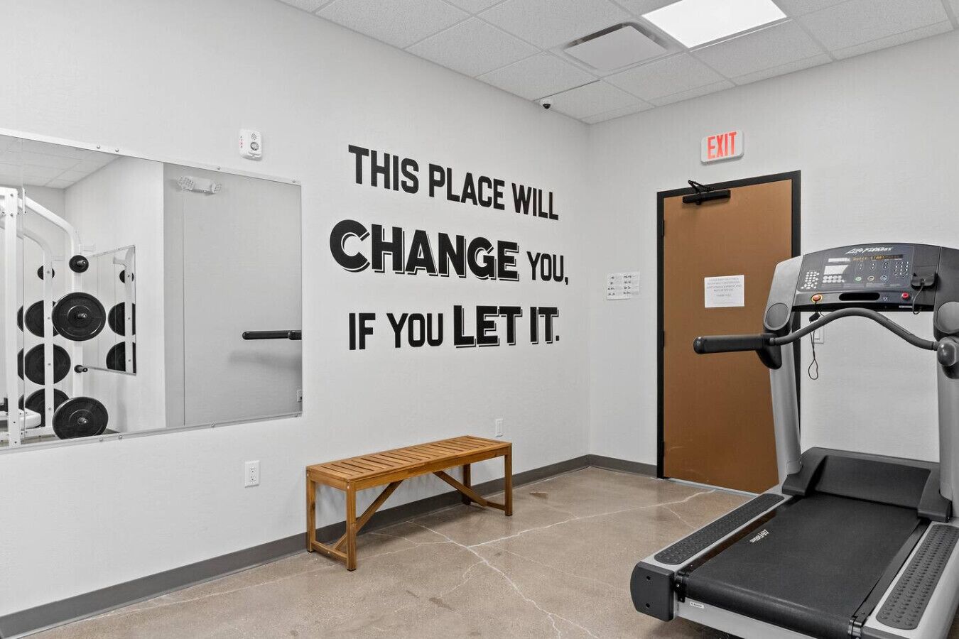 Un gimnasio con una frase motivacional en la pared, una cinta de correr y un banco.