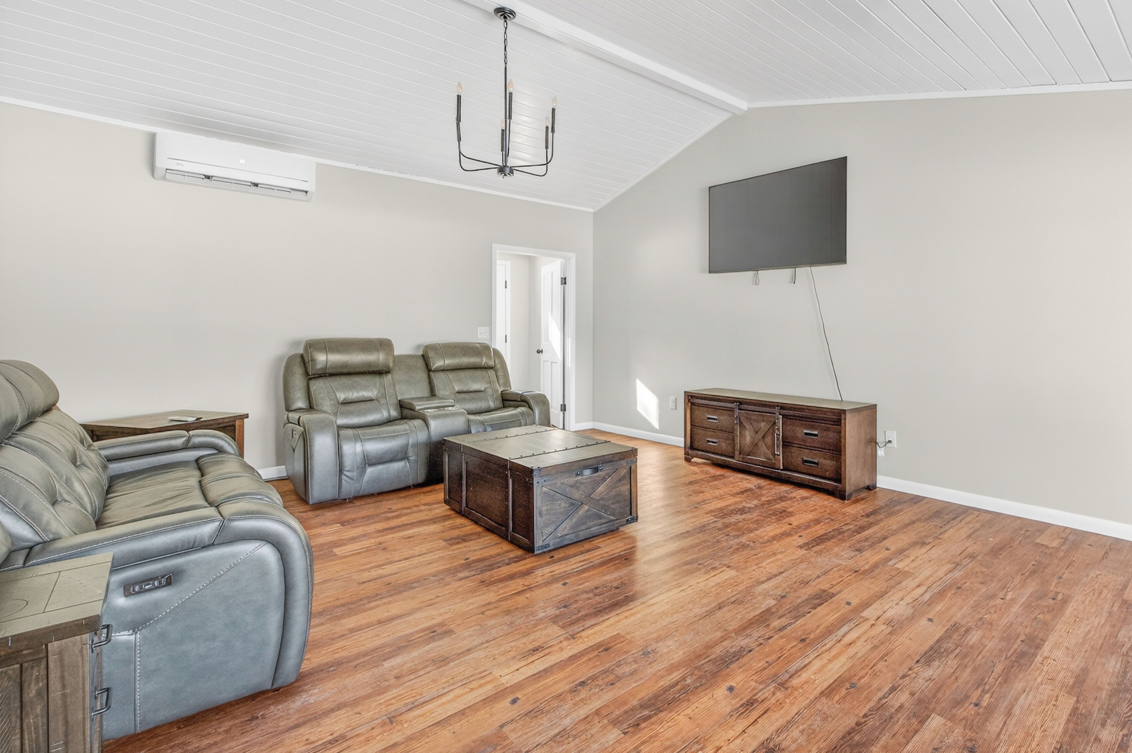 Een ruime woonkamer met een grote grijze hoekbank, een houten salontafel, een donker houten kast en een wandgemonteerde flatscreen televisie.