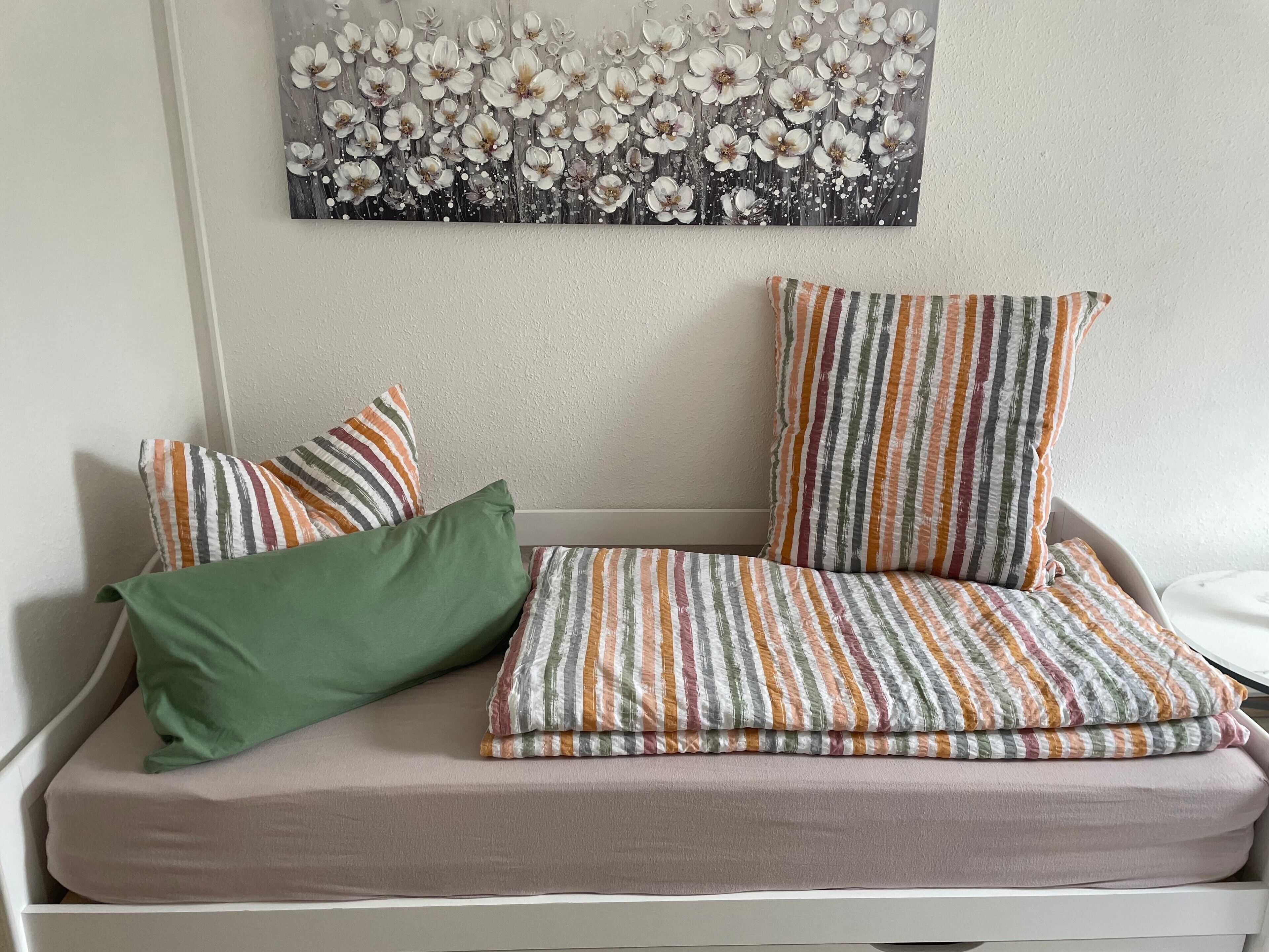 A bed with a striped pillow, a solid green pillow, and a floral artwork above.