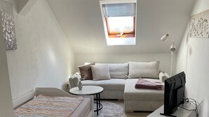 Quarto estilo loft com teto inclinado, uma janela pequena, um sofá-cama, uma TV em um suporte e uma mesinha de cabeceira.