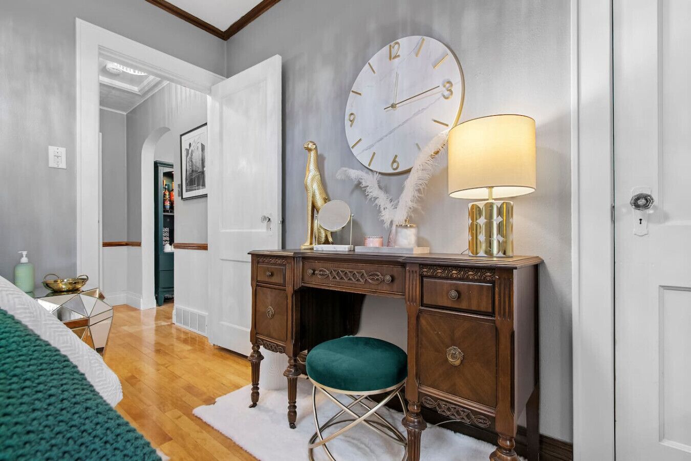 Une chambre à coucher avec une commode en bois, un fauteuil vert, un grand miroir rond, une horloge murale et une lampe de chevet.