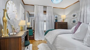 Ein Schlafzimmer mit einem Bett, einer Kommode, einem Schreibtisch, einem Stuhl, einer Lampe, einem Kronleuchter und Vorhängen.