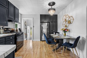 A modern kitchen with dark cabinets, a stainless steel refrigerator, a dining table with blue chairs, and a chandelier. - AfterWIRTZ - Walking distance to Lambeau/Resch Ctr (Green Bay)