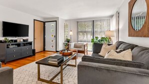 A modern living room with a flat-screen TV, dark furniture, a coffee table, and a large mirror. - Biemeret Garden - Steps from Lambeau, Resch Center (Green Bay)