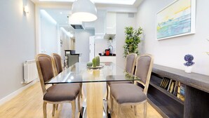 Een moderne eetruimte met een glazen tafel, houten stoelen, een witte hanglamp, een boekenkast en een schilderij aan de muur.
