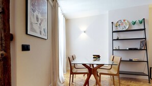 Een eetruimte met een glazen tafel, houten stoelen, een wandlamp en een boekenplank met decoratieve objecten.