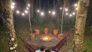 A hammock between trees with a fire pit in the center, surrounded by string lights. - villaggio farnese (Campos do Jordão)