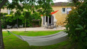 A hammock in a grassy area with a stone building, outdoor seating, and a pergola with climbing plants.