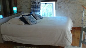 Una cama con ropa blanca y almohadas estampadas en azul, suelo de madera, ventana con vista a la vegetación y una pared con un tapiz de gallo.