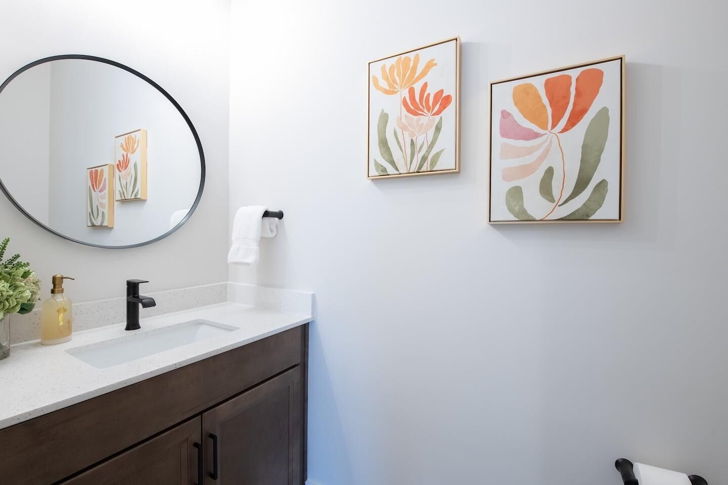 Uma casa de banho com um espelho redondo, uma sanita branca, armários escuros e duas obras de arte floral emolduradas na parede.
