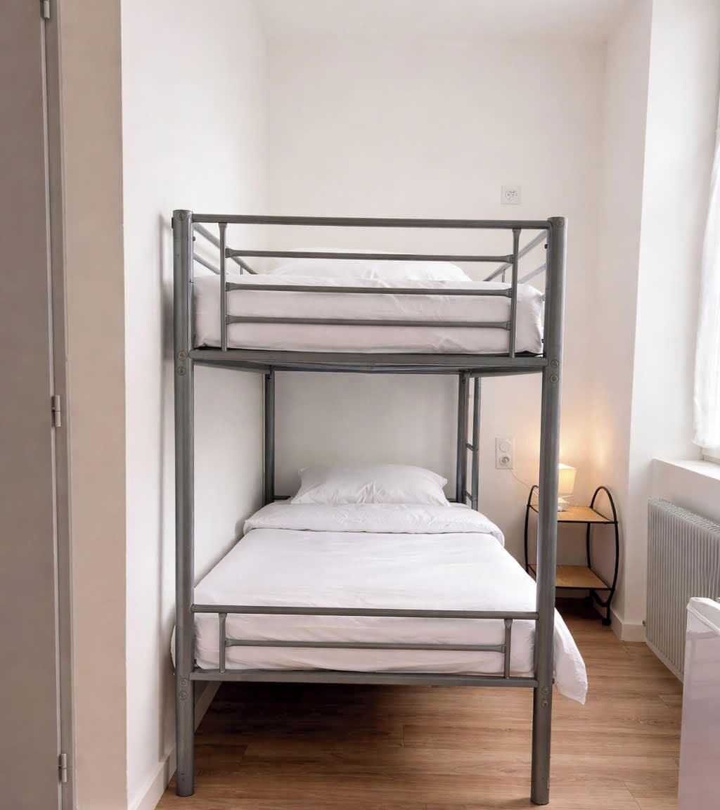 Una cama individual superpuesta en una habitación con un radiador, una mesita pequeña con una lámpara y una puerta.