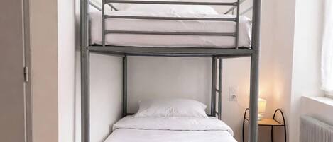 Una cama individual superpuesta en una habitación con un radiador, una mesita pequeña con una lámpara y una puerta.
