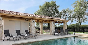 Un espace aménagé au bord de la piscine, avec des chaises longues, un coin salon abrité et un bâtiment en pierre surmonté d’un toit en tuiles.