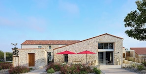 Un bâtiment en pierre avec un toit recouvert de tuiles, une grande porte vitrée et des parasols rouges dans le hall d’entrée.