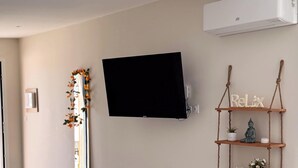 A living room with a flat-screen TV mounted on the wall, a wooden shelf with decorative items, and a mirror with a floral garland. - Tranquiloue - Appart 4 pers - Puy du Fou 17 min (Chanverrie)