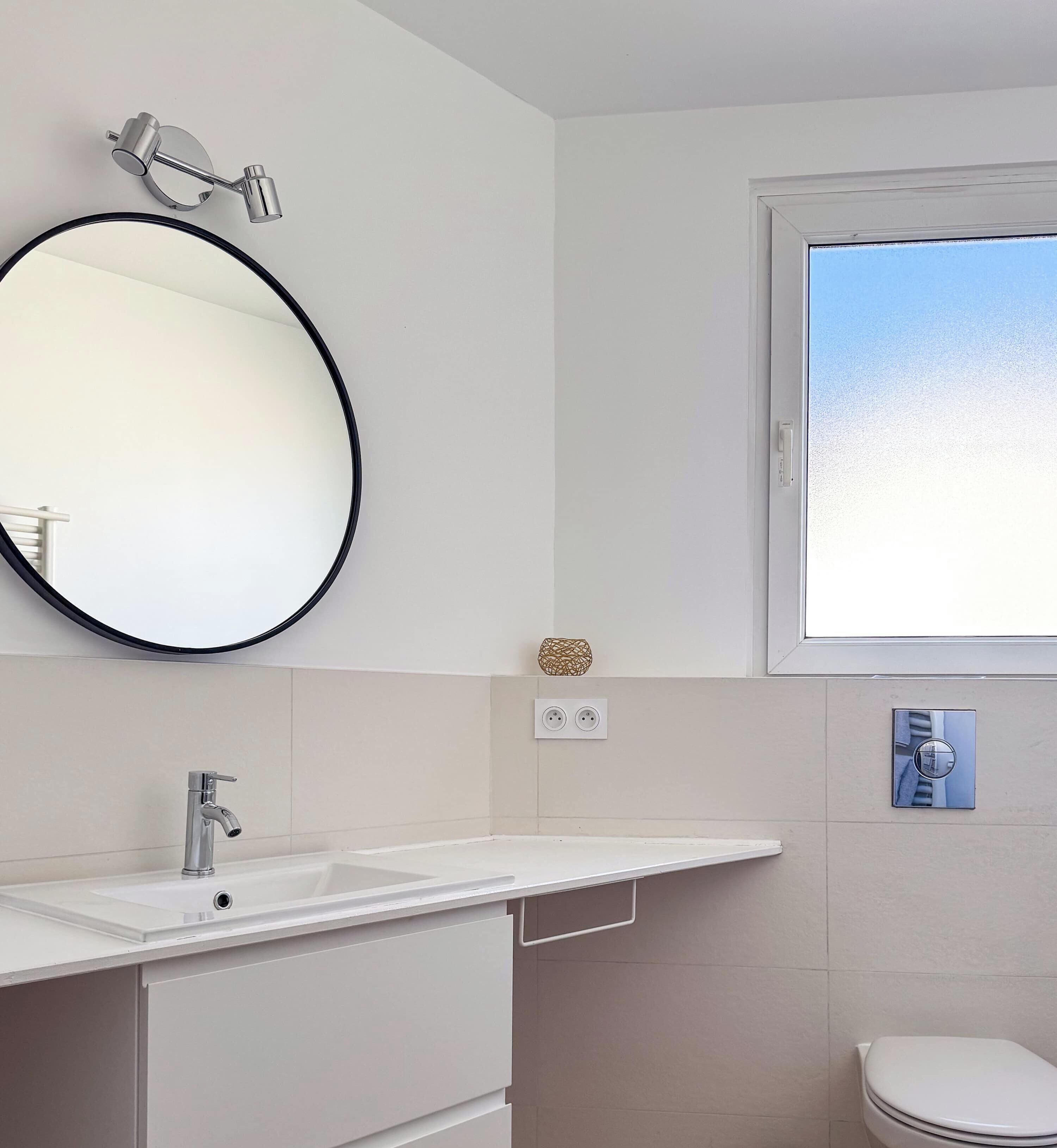 Un baño con un espejo redondo, un lavabo blanco, un inodoro y una ventana.