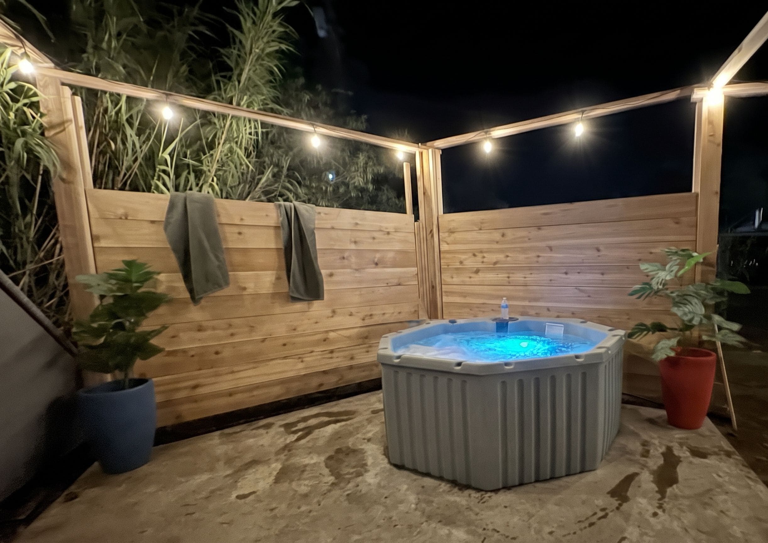 An outdoor hot tub area with a wooden fence, potted plants, and string lights.