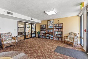 A library with bookshelves, two wicker chairs, a patterned carpet, and a large window. - Sherwin Condominium 501 (Daytona Beach Shores)