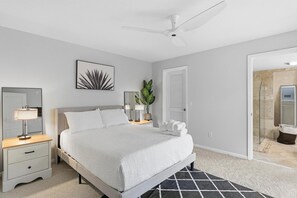 Ein Schlafzimmer mit einem Bett, Nachttischen, einem Deckenventilator, einer gerahmten Kunstwerk, einer Topfpflanze und einer Badezimmertür.