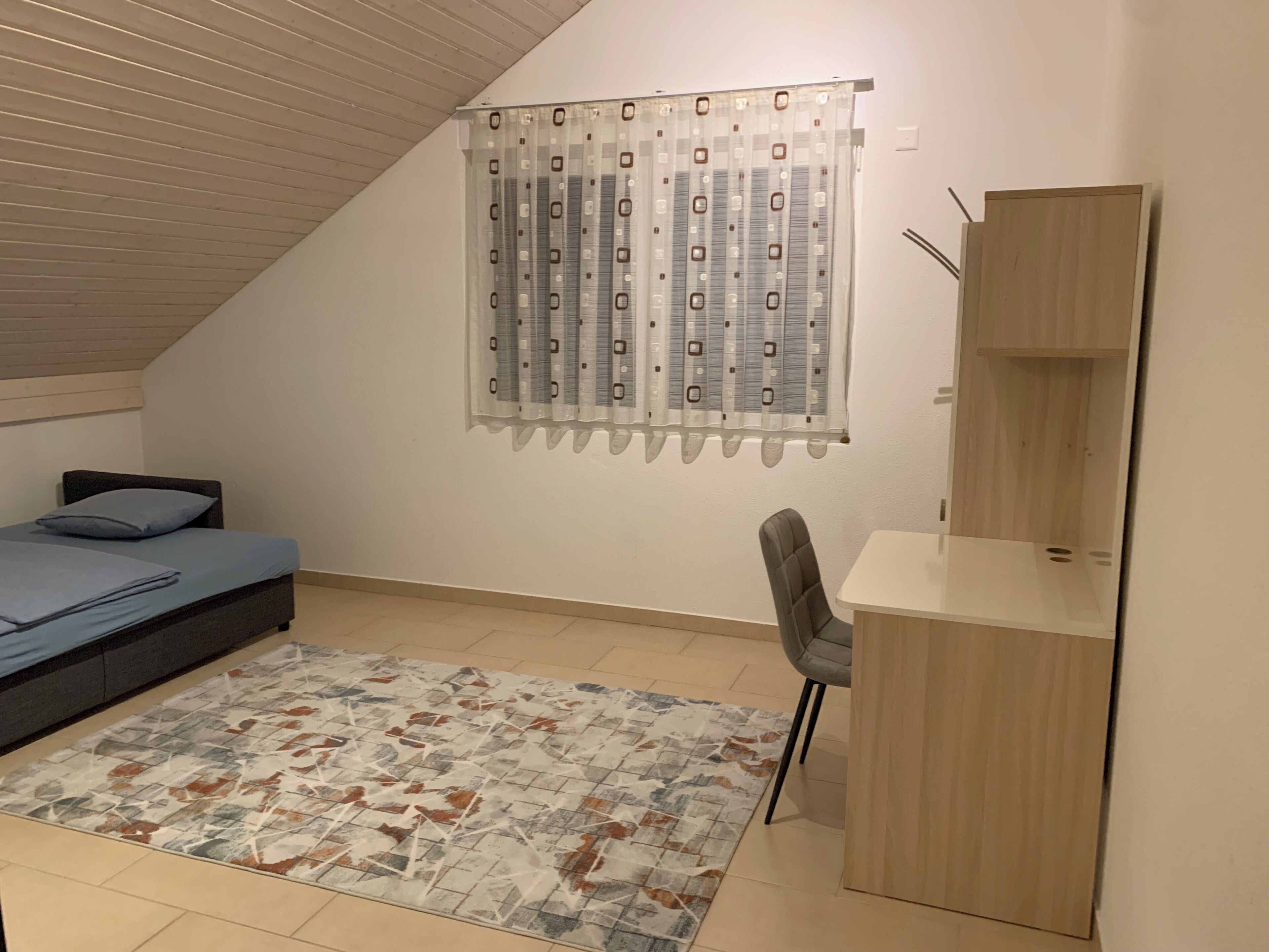 A bedroom with a bed, a desk, a chair, a patterned rug, and a window with curtains.